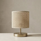 Table lamp with natural fabric cylinder shade and brass finish metal base, warm diffused light